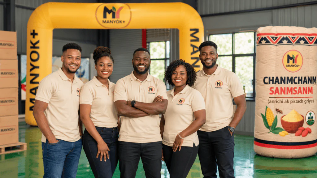 Manyok Plus team standing in a warehouse with Haitian products and branded inflatable displays.
